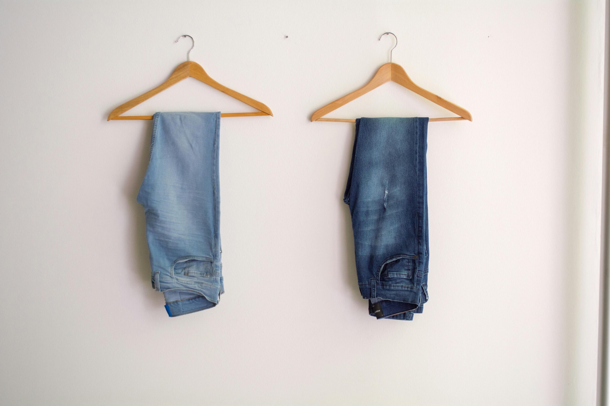 Two pairs of blue denim jeans neatly hanging on wooden hangers against a plain wall.